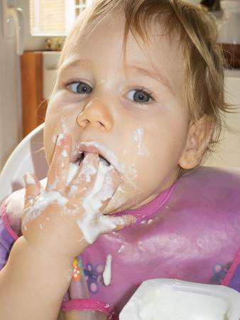 blonde caucasian baby 1 year eating natural yogurt with her hand in high-chair kitchenの写真素材