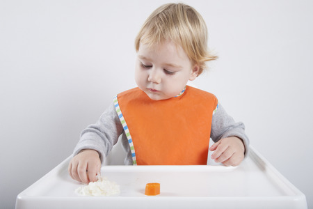 blonde caucasian baby seventeen month age orange bib grey sweater eating meal with her hand in white high-chairの写真素材