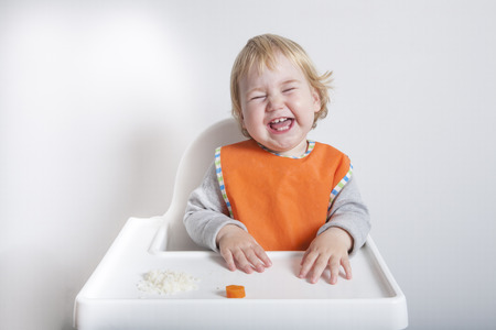 blonde caucasian baby seventeen month age orange bib grey sweater eating rice carrot on white high-chair laughing and smilingの写真素材