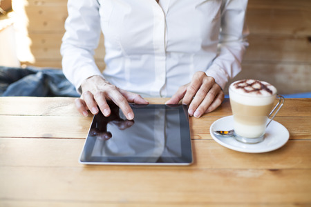 woman touching digital tablet blank screen with cappuccino coffee cup on light brown wooden table cafeの写真素材