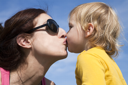 two years old age blonde baby yellow shirt kissing in mouth to brunette woman mother sunglasses with blue sky backgroundの写真素材