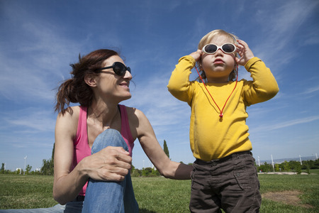 two years aged blonde happy baby yellow shirt with white kid sunglasses next to brunette woman mother smiling sitting on green grass lawn in parkの写真素材