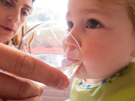 face of baby two years old with green bib drinking water from glass crystal cup holding by woman mother handの写真素材