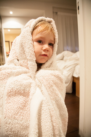 blonde caucasian baby two years old age chubby face wrapped in white bath towel looking indoor bedroom homeの写真素材