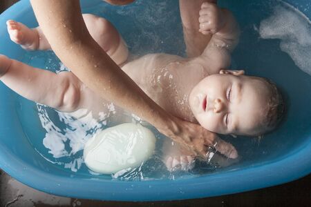 six months age blonde baby body and face washing by woman mother hands in blue little plastic bath indoor with brown backgroundの写真素材