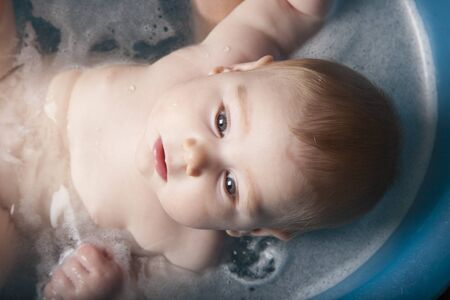 six months age blonde baby body and face washing by woman mother hands in blue little plastic bath indoor with brown backgroundの写真素材