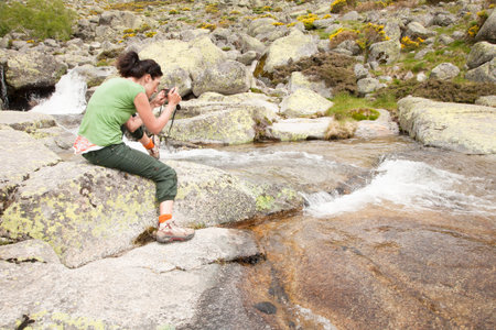 brunette tourist woman with green shirt and trousers sitting on rock with camera photo, as...