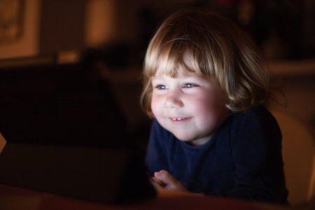 blonde two years old baby blue sweater, sitting inside home at night reading and watching digital...