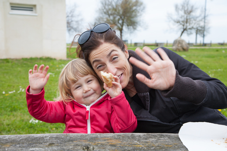 little blonde cute child sitting with woman eating bread piece and looking and greeting with hand in park green grass backgroundの写真素材