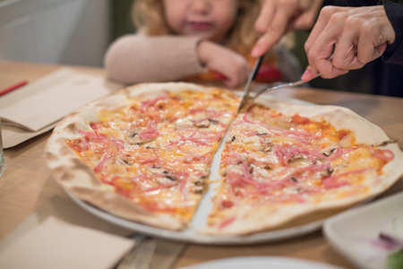 Three years old blonde child at restaurant table looking woman hands cutting ham and cheese pizza with fork and knife, dividing by halfの写真素材