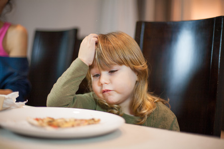 Three years old blonde child looking at pizza piece in her dish with expression she does not like it, sitting in dark brown chair, in table with beige tablecloth at home, next to motherの写真素材