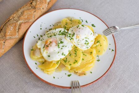 aerial view of white circular dish with typical Spanish food, sliced fried potatoes and eggs and chopped parsley, with two silver forks and bread on beige tableclothの写真素材