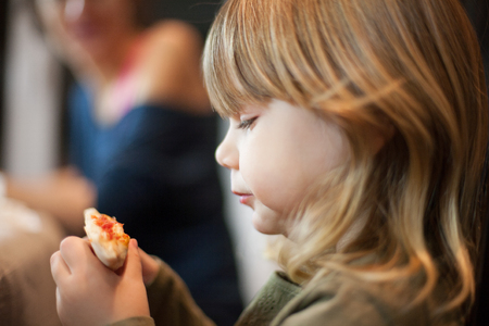 Three years old blonde hungry child eating with her hands and looking edge of pizza portion, with woman in backgroundの写真素材