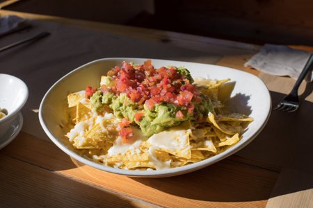 white big bowl with nachos and guacamole, chopped tomato and cheese, on wooden table at restaurantの写真素材