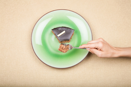aerial view hand with fork taking piece of triangular portion of chocolate cake on green circular dish, on golden backgroundの写真素材
