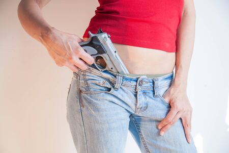 detail close up of plastic toy silver false pistol gun holding in jeans waist of woman with red tee shirtの写真素材