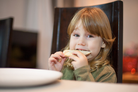 Three years old blonde hungry child biting pizza piece in her hands, sitting in dark brown chair, in table with grey tablecloth at home, looking at cameraの写真素材