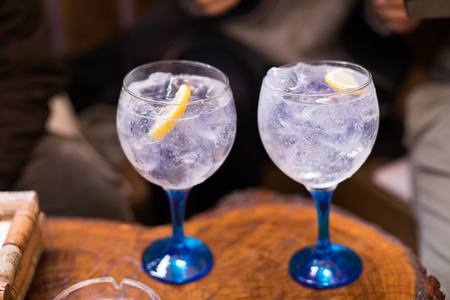 two transparent glass wide cups, with blue crystal, full of gin tonic cocktail drink, with lemon slice and ice cubes, on wooden tableの写真素材