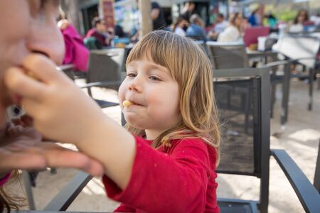 blonde three years old child, with red shirt, feeding mother with cheese puff, sitting in terrace exterior bar cafe with grey chairの写真素材