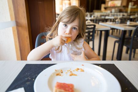 portrait of blonde caucasian child three years old with white shirt, looking with plastic water bottle in hands, with a slice of bread with tomato on dish, sitting indoor in restaurantの写真素材