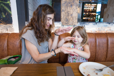 Four years age blonde happy girl playing with hands with woman mother and smiling sitting in brown leather sofa at restaurantの写真素材