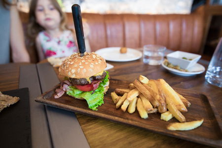 still life of beef burger, with cheese, bacon, lettuce and tomato slice, inside bun bread with seeds and potatoes french fries on brown wooden tray on table at cafe restaurant. Little girl lookingの写真素材