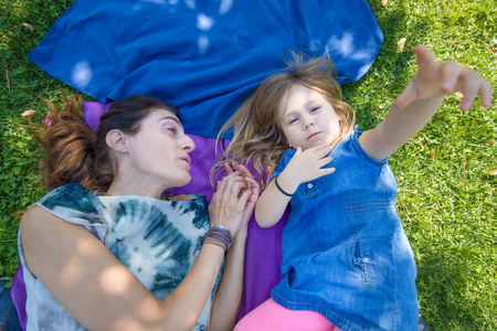 Four years old blonde child playing with hand, lying with woman mother on towels in the green grass of parkの写真素材