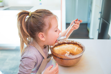 hungry four years old girl tasting or eating chocolate cream with spoon from glass bowl, at kitchen of homeの写真素材