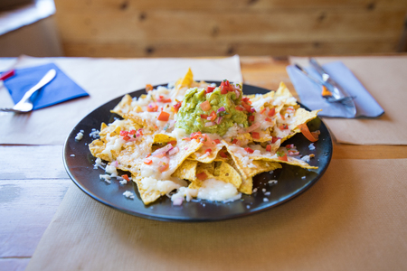 black plate with nachos and guacamole, chopped tomato and cheese, on wooden table with paper placemats at restaurantの写真素材