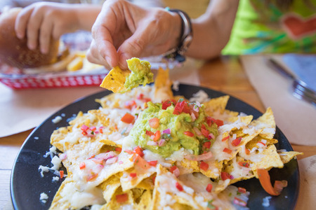 woman hand dipping nacho chips in avocado guacamole, cheese and chopped tomato on black plate, on wooden table with paper placemats at restaurantの写真素材