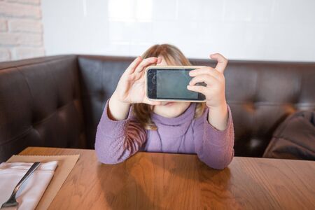 Portrait of four years old child with violet jersey siting at a restaurant table, with a smartphone mobile in her hands, hiding her eyes, and showing blank screenの写真素材