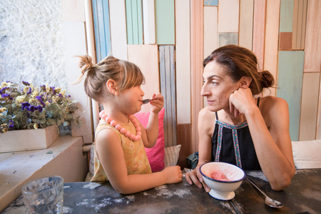 Three years old blonde girl with yellow dress an necklace eating strawberry ice cream with spoon from bowl, looking at woman mother, sitting indoor in restaurantの写真素材