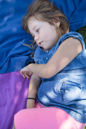 four years old blonde girl lying on towels in the green grass of park looking sideway with thoughtful or imagining faceの写真素材
