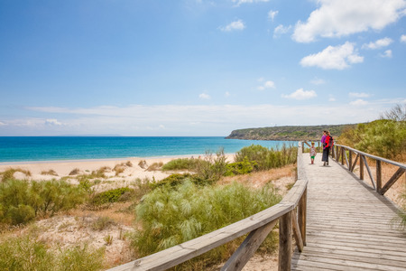 landscape of Beach Bolonia in Tarifa, Cadiz, Andalusia, Spain. Four years old blonde girl with woman mother with backpack walking on wooden footbridgeの写真素材