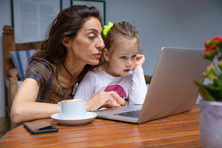 mother teaching her little daughter, girl four years old sitting on mother legs, surfing internet in laptop pc computer together, with coffee cup and black mobile smart phone on brown wooden tableの写真素材