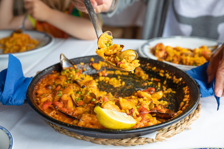 serving typical Spanish paella with seafood and seashell (mussel, squid, clam), rice and lemon with big spoon on white tablecloth of restaurantの写真素材