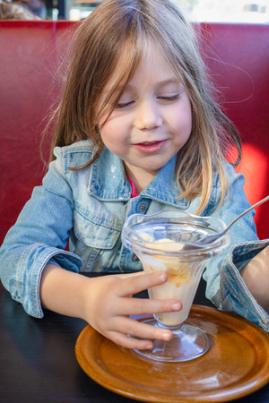 Portrait of five years old blonde little child with blue denim jacket, sitting in red sofa and black table in a restaurant, eating vanilla ice cream with spoon from crystal cup. vertical shotの写真素材