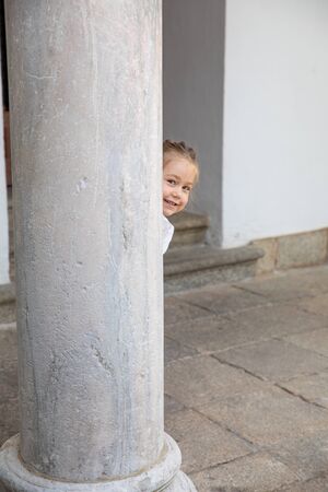 little child four years old peeking face and looking at smiling behind ancient marble columnの写真素材