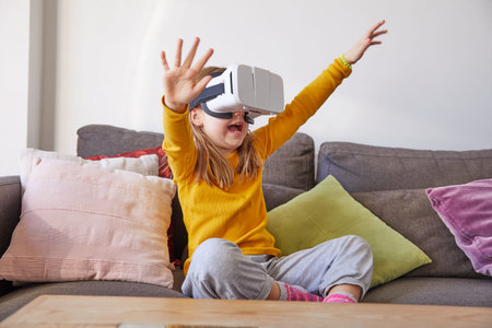 six years old child sitting on a brown sofa at home watching virtual reality headset glasses...