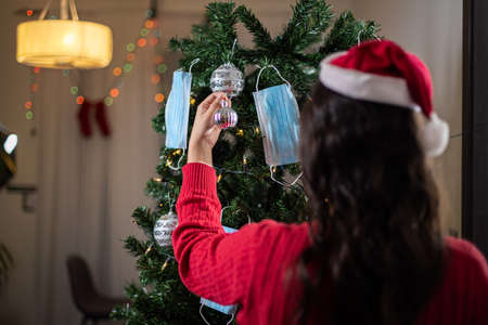 Young woman in santa hat decorating christmas tree with medical masks during coronavirus pandemicの写真素材