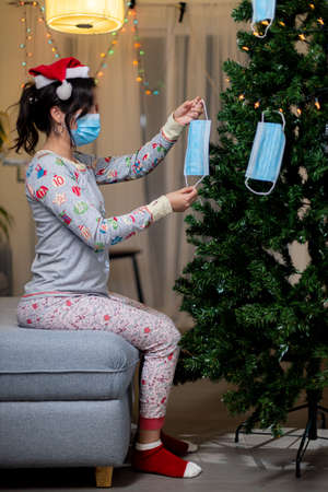 Young woman in Santa Claus hat and pajamas decorating Christmas tree with medical masks.の写真素材