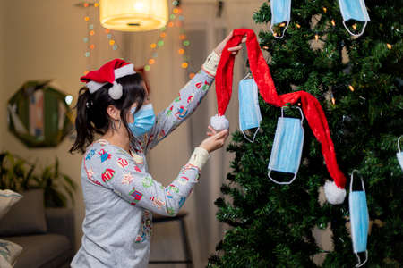 Young woman in Santa Claus hat and pajamas decorating the Christmas tree with medical masks.の写真素材
