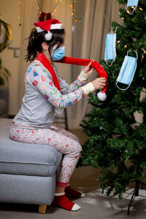 Girl in a medical mask and a red sweater puts on gloves on a Christmas tree.の写真素材