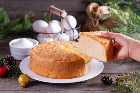 Homemade sponge cake on a white plate on a wooden table with Christmas tree branches, cones and...