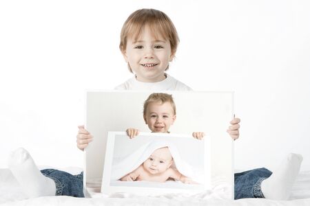A cute young boy, sits on the floor, holding baby photographs of himself. The photographs are arranged such that the boy is holding a photo of his younger self, holding a photo of his baby self. Signifies growth.の写真素材