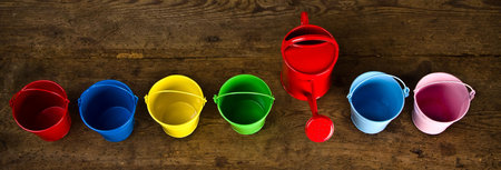 Six coloured buckets in line with red watering can on timber floor taken by top viewの写真素材