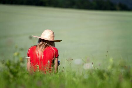 A girl sitting in a field in summer wearing a straw sun hat and red t.shirt. の写真素材