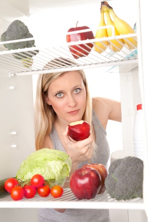 Attractive female teenager eating apple and looking at healthy fruit and vegetables in refrigerator.の写真素材