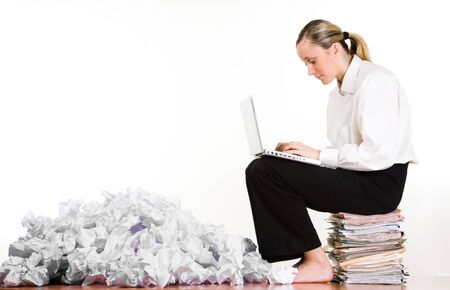 A woman using a laptop computer, sitting on a stack of file folders and paperwork beside a pile of crumpled paper.  White backgroundの写真素材