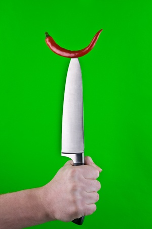 A view of a hand holding a large butcher knife vertical with a single red pepper on its sharp point.  Green background.の写真素材